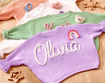 Op de afbeelding: Vier pastelkleurige gebreide truien worden getoond, elk met een unieke naam of initiaal. De truien zijn in tinten perzik, wit, groen en lavendel. De lavendelkleurige trui heeft de naam "Olivia" in witte letters.