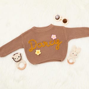 Op de afbeelding: Een bruine gebreide trui met de naam "Darcy" geborduurd in geel garen. De trui is versierd met drie kleine gehaakte bloemen in roze, geel en wit.