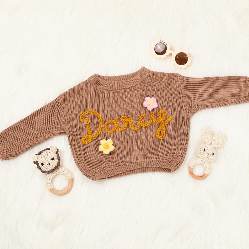 Op de afbeelding: Een bruine gebreide trui met de naam "Darcy" geborduurd in geel garen. De trui is versierd met drie kleine gehaakte bloemen in roze, geel en wit.