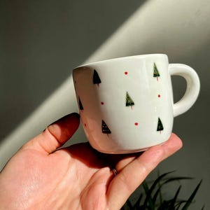 Könnte beinhalten: Weiße Keramik-Tasse mit grünen Weihnachtsbäumen und roten Punkten verziert. Die Tasse hat einen kleinen Henkel und wird in einer Hand gehalten. Die Tasse ist ein festlicher Artikel, perfekt für Weihnachtsgetränke.