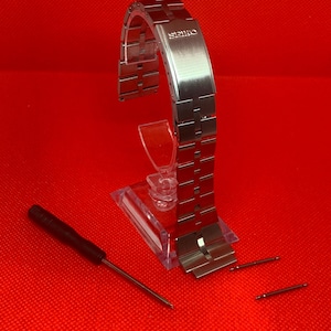 Peut inclure: Un bracelet de montre argenté avec le mot "SEIKO" gravé sur le fermoir. Le bracelet est sur un présentoir transparent avec deux petits barillets argentés et un petit tournevis noir.