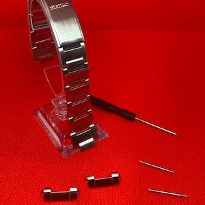 Puede incluir: Una correa de reloj plateada con la palabra "Seiko" grabada en ella. La correa está en un expositor transparente. Hay dos eslabones de correa de reloj plateados y dos pasadores de resorte plateados sobre una superficie roja.