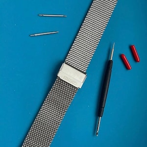 May include: A silver metal watch band with a mesh design. The band has the word "SEIKO" engraved on the clasp. The band is laying on a blue surface with two small silver metal pins and a black tool.