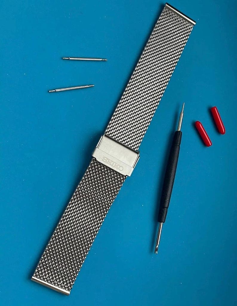 May include: A silver metal watch band with a mesh design. The band has the word "SEIKO" engraved on the clasp. The band is laying on a blue surface with two small silver metal pins and a black tool.