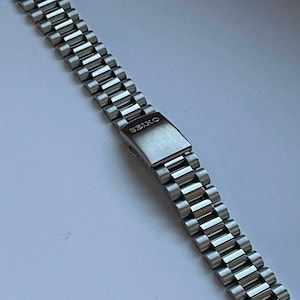 May include: A silver-toned stainless steel watch bracelet featuring a multi-link design and a fold-over clasp. The clasp is engraved with the brand name SEIKO in capital letters. The metal links have a brushed finish, creating a uniform appearance.