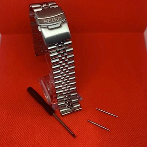 May include: A silver watch band with the word "SEIKO" engraved on the clasp. The band is on a red surface with a screwdriver and two spring bars.