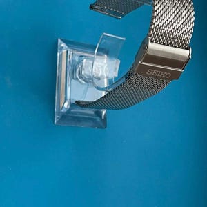 May include: A silver metal watch band with a mesh design. The band is on a clear plastic display stand. The band has the word "SEIKO" engraved on the clasp.