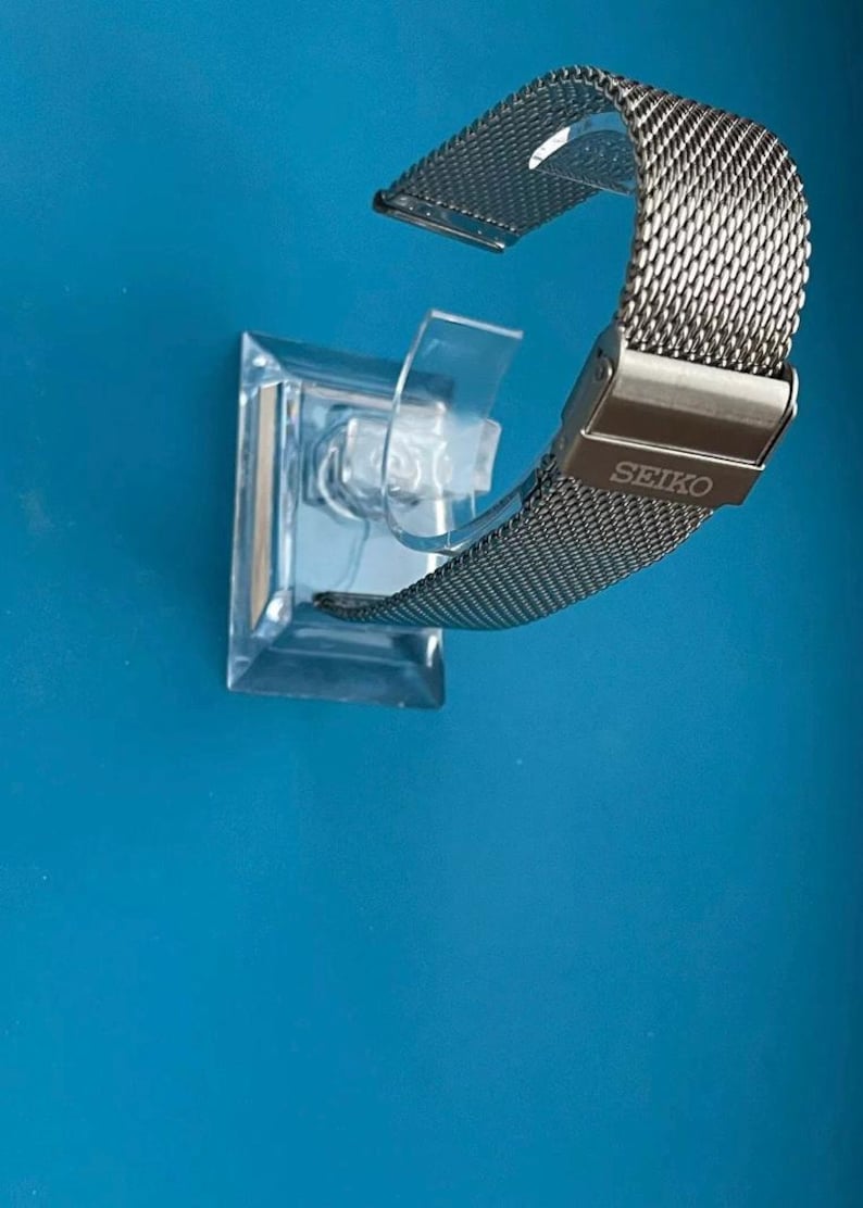 May include: A silver metal watch band with a mesh design. The band is on a clear plastic display stand. The band has the word "SEIKO" engraved on the clasp.