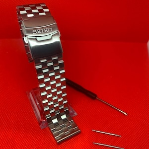 Puede incluir: Una correa de reloj de metal plateado con la palabra "SEIKO" grabada en el cierre. La correa está sobre una superficie roja con dos pasadores de reloj de metal plateado.