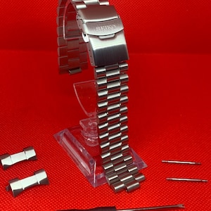 Può includere: Un cinturino per orologio Seiko in acciaio inossidabile con chiusura deployante, esposto su un supporto trasparente su sfondo rosso. Include due maglie di ricambio, due perni e un utensile per orologi. Il cinturino è composto da maglie rettangolari.