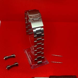 May include: A silver watch band with a clasp that says "SEIKO" on it. The band is on a clear stand. There are two silver watch band links and a small screwdriver on a red surface.