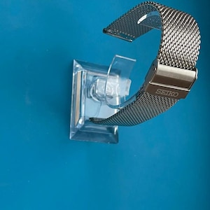 May include: A silver metal watch band with a mesh design. The band is on a clear plastic display stand. The band has the word "SEIKO" printed on it.