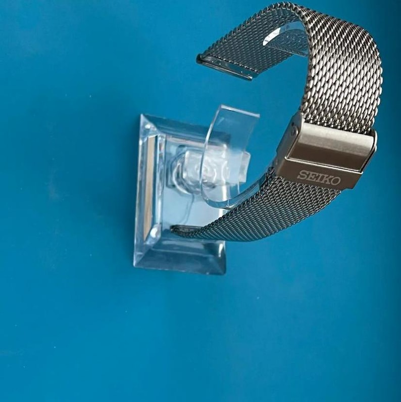 May include: A silver metal watch band with a mesh design. The band is on a clear plastic display stand. The band has the word "SEIKO" printed on it.