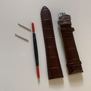 Puede incluir: Una correa de reloj de cuero marrón con una hebilla plateada que dice "SEIKO". La correa viene con dos pasadores de resorte y una herramienta para quitar los pasadores de resorte.