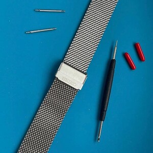 May include: A silver metal watch band with a mesh design. The band has a clasp with the word "SEIKO" engraved on it. The band is laying on a blue surface with two small silver metal pins and a black tool.
