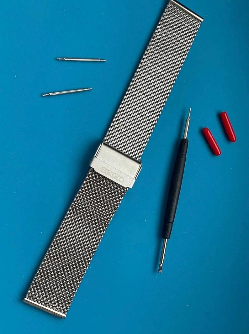 May include: A silver metal watch band with a mesh design. The band has a clasp with the word "SEIKO" engraved on it. The band is laying on a blue surface with two small silver metal pins and a black tool.