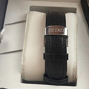 Puede incluir: Una correa de reloj de cuero negro con una hebilla plateada que dice "SEIKO".