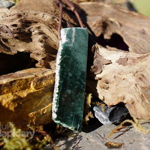 Peut inclure: Un pendentif rectangulaire en pierre vert foncé avec des motifs tachetés blancs et un bord latéral brut et non poli. Le pendentif est suspendu à un cordon marron fin et porte le texte luki lapidary artisan lapidary dans le coin inférieur gauche.