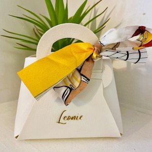 Op de afbeelding: Een witte handtasvormige geschenkdoos met een handvat en een gele strik. De doos heeft de naam "Leonie" in gouden letters op de voorkant gedrukt. Een sjaal met patroon is aan het handvat vastgebonden.