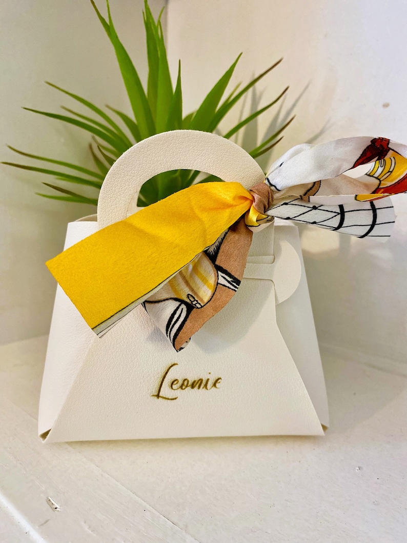 Op de afbeelding: Een witte handtasvormige geschenkdoos met een handvat en een gele strik. De doos heeft de naam "Leonie" in gouden letters op de voorkant gedrukt. Een sjaal met patroon is aan het handvat vastgebonden.