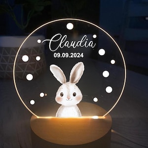 K&ouml;nnte beinhalten: Personalisiertes Nachtlicht mit einer Hasenillustration. Die transparente Acrylscheibe zeigt den Namen "Claudia" und das Datum 09.09.2024 in wei&szlig;er Schrift. Das Licht steht auf einem Holzsockel und erzeugt ein warmes Leuchten.