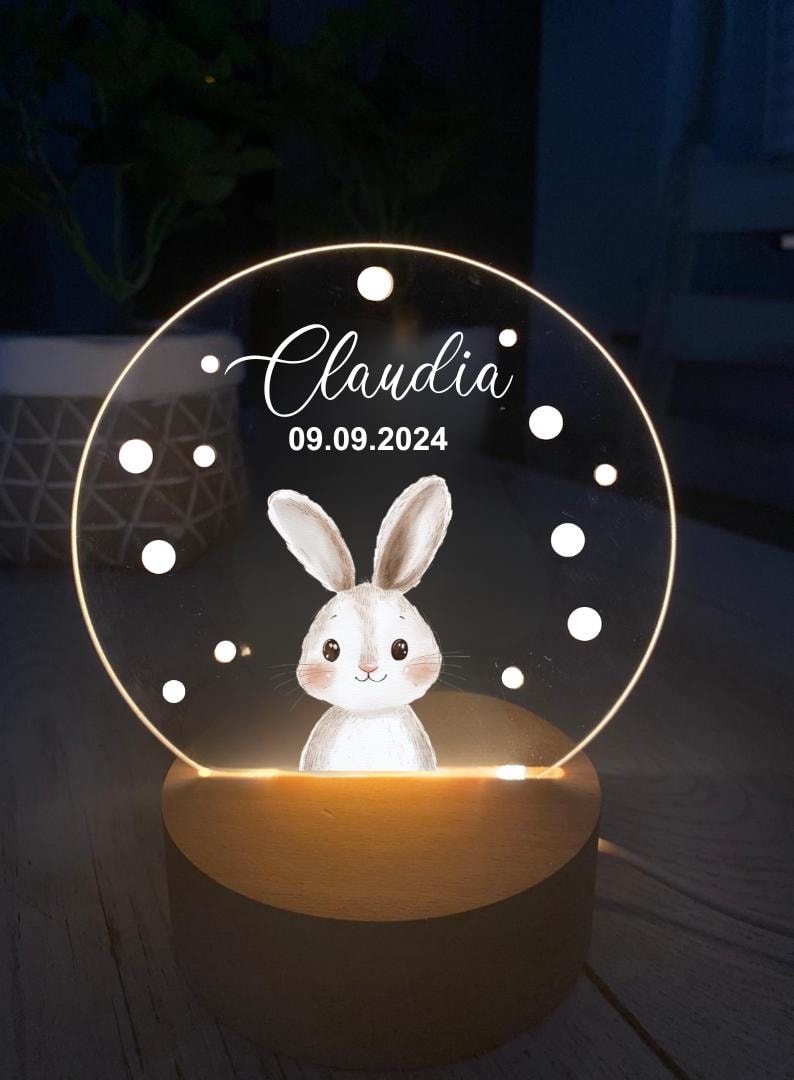 K&ouml;nnte beinhalten: Personalisiertes Nachtlicht mit einer Hasenillustration. Die transparente Acrylscheibe zeigt den Namen "Claudia" und das Datum 09.09.2024 in wei&szlig;er Schrift. Das Licht steht auf einem Holzsockel und erzeugt ein warmes Leuchten.