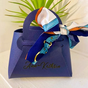 Op de afbeelding: Een marineblauwe geschenktas met een handvat, met de naam "Ann-Kathrin" in gouden script. Een sjaal met patronen in wit, blauw en oranje is aan het handvat geknoopt. De tas staat voor een groene plant.