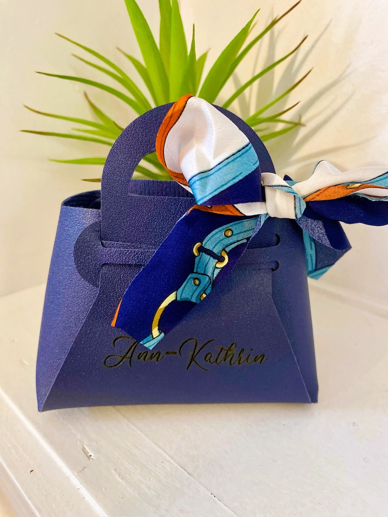 Op de afbeelding: Een marineblauwe geschenktas met een handvat, met de naam "Ann-Kathrin" in gouden script. Een sjaal met patronen in wit, blauw en oranje is aan het handvat geknoopt. De tas staat voor een groene plant.