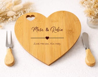 Op de afbeelding: Een hartvormige houten snijplank met de namen "Mara & Robin" en de zin "Zwei Herzen. Ein Weg." gegraveerd. Een klein hartje staat tussen de namen. Een kaasvork en mes met houten handvatten staan aan weerszijden.