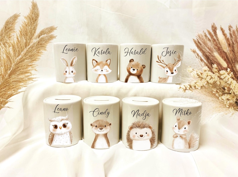 May include: Eight ceramic coin banks with animal illustrations, including a rabbit, fox, bear, deer, owl, otter, hedgehog, and squirrel. Each bank has a name printed on it in black script. The banks are light beige.