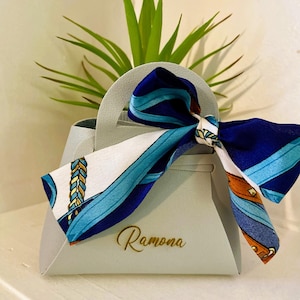 Op de afbeelding: Een lichtgrijze handtasvormige geschenkdoos met een handvat, vastgebonden met een sjaal met patronen in blauw, wit en bruin. De naam "Ramona" staat in gouden letters op de voorkant. Een groene plant staat op de achtergrond.