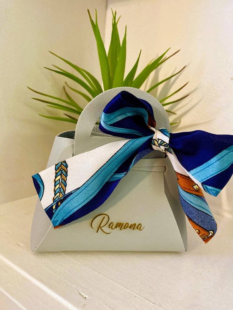 Op de afbeelding: Een lichtgrijze handtasvormige geschenkdoos met een handvat, vastgebonden met een sjaal met patronen in blauw, wit en bruin. De naam "Ramona" staat in gouden letters op de voorkant. Een groene plant staat op de achtergrond.