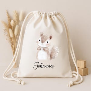 Op de afbeelding: Een cr&egrave;mekleurige rugzak met trekkoord met een aquarel illustratie van een schattig eekhoorntje. Het eekhoorntje is in bruin- en beigetinten, met een pluizige staart. De naam "Johannes" staat eronder in cursief.