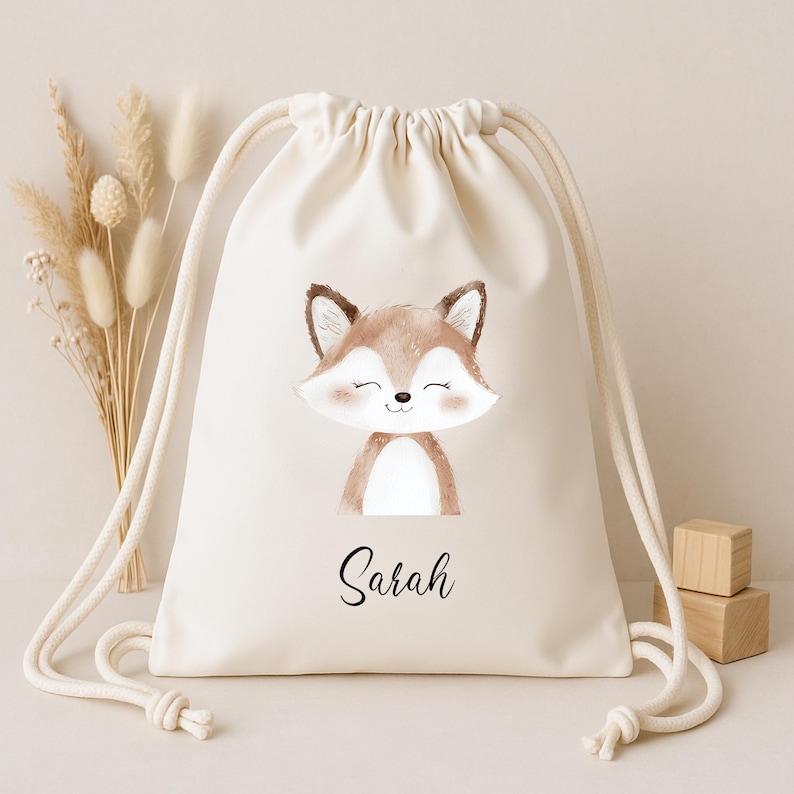 Op de afbeelding: Een cr&egrave;mekleurige rugzak met trekkoord met een aquarel illustratie van een lachende vos met bruine en witte vacht. De naam "Sarah" staat in zwart schrift eronder. De tas heeft witte trekkoorden en is geschikt voor het dragen van spullen.