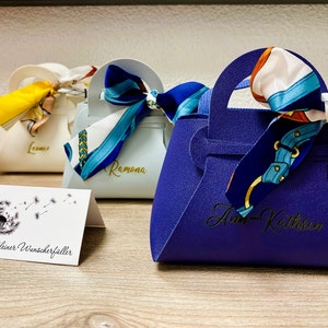 Op de afbeelding: Een verzameling kleine, handtasvormige geschenkdozen in verschillende kleuren, elk versierd met een sjaal met patroon die aan het handvat is vastgebonden. De dozen hebben namen erop gedrukt. Een kaart met een paardenbloemillustratie staat op de voorgrond.