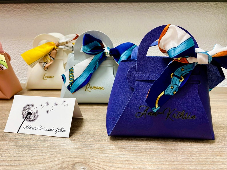 Op de afbeelding: Een verzameling kleine, handtasvormige geschenkdozen in verschillende kleuren, elk versierd met een sjaal met patroon die aan het handvat is vastgebonden. De dozen hebben namen erop gedrukt. Een kaart met een paardenbloemillustratie staat op de voorgrond.