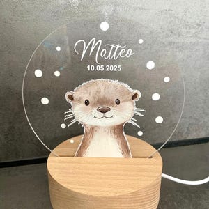 K&ouml;nnte beinhalten: Dekoratives Nachtlicht mit einer Otter-Illustration. Die transparente Acrylplatte tr&auml;gt den Namen "Matteo" und das Datum "10.05.2025". Das Licht steht auf einem runden Holzsockel. Das Design umfasst wei&szlig;e Punkte.
