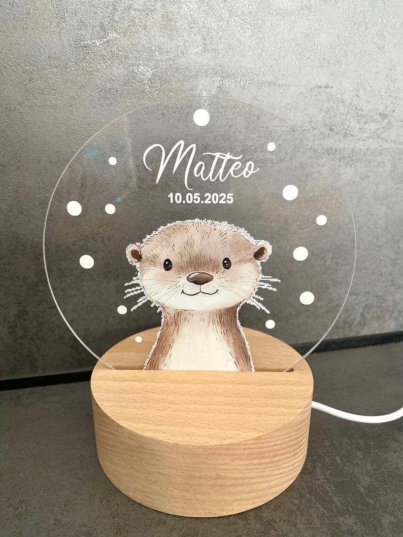 K&ouml;nnte beinhalten: Dekoratives Nachtlicht mit einer Otter-Illustration. Die transparente Acrylplatte tr&auml;gt den Namen "Matteo" und das Datum "10.05.2025". Das Licht steht auf einem runden Holzsockel. Das Design umfasst wei&szlig;e Punkte.