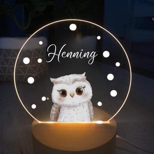 K&ouml;nnte beinhalten: Dekoratives Nachtlicht mit einer Eulenillustration. Der Name "Henning" steht &uuml;ber dem Bild. Die Eule befindet sich auf einer transparenten, kreisf&ouml;rmigen Acrylplatte mit einem Holzsockel. Das Licht strahlt warm.