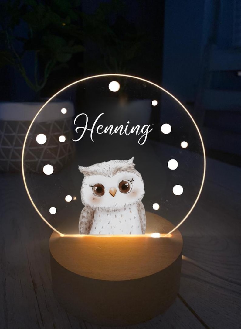 K&ouml;nnte beinhalten: Dekoratives Nachtlicht mit einer Eulenillustration. Der Name "Henning" steht &uuml;ber dem Bild. Die Eule befindet sich auf einer transparenten, kreisf&ouml;rmigen Acrylplatte mit einem Holzsockel. Das Licht strahlt warm.