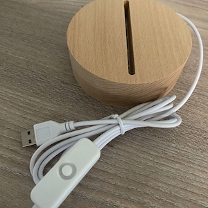 K&ouml;nnte beinhalten: Ein runder, hellfarbiger Holzsockel mit einem vertikalen Schlitz, verbunden mit einem wei&szlig;en USB-Kabel und einem wei&szlig;en Inline-Ein/Aus-Schalter. Der Sockel ist f&uuml;r eine dekorative Lampe.