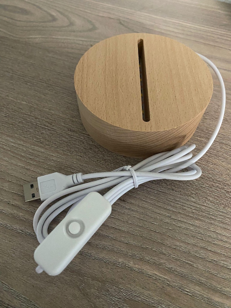 K&ouml;nnte beinhalten: Ein runder, hellfarbiger Holzsockel mit einem vertikalen Schlitz, verbunden mit einem wei&szlig;en USB-Kabel und einem wei&szlig;en Inline-Ein/Aus-Schalter. Der Sockel ist f&uuml;r eine dekorative Lampe.