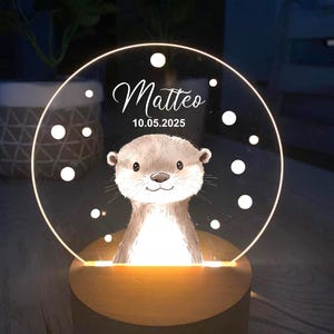 K&ouml;nnte beinhalten: Personalisiertes Nachtlicht mit einer Otter-Illustration. Die transparente Acrylscheibe wird von einem warmen Licht beleuchtet und zeigt den Namen "Matteo" und das Datum "10.05.2025". Der Sockel ist aus Holz.