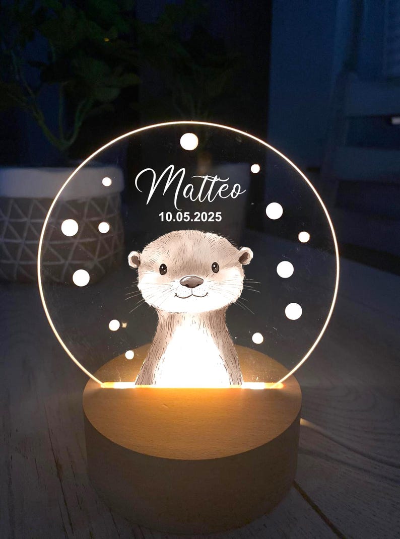K&ouml;nnte beinhalten: Personalisiertes Nachtlicht mit einer Otter-Illustration. Die transparente Acrylscheibe wird von einem warmen Licht beleuchtet und zeigt den Namen "Matteo" und das Datum "10.05.2025". Der Sockel ist aus Holz.