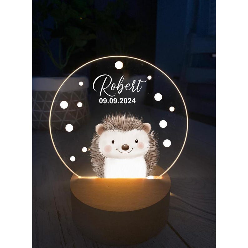K&ouml;nnte beinhalten: Ein dekoratives Nachtlicht mit einer Illustration eines l&auml;chelnden Igels. Das klare, kreisf&ouml;rmige Licht tr&auml;gt den Namen "Robert" und das Datum "09.09.2024". Das Licht steht auf einem Holzsockel und strahlt warmes Licht aus.