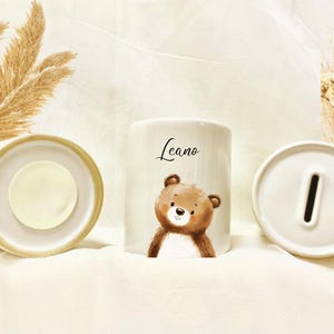 May include: White ceramic piggy bank with a cute brown bear illustration and the name "Leano" in script. The bank is cylindrical with a slot for coins on top and a removable base. Decorative dried plants are in the background.