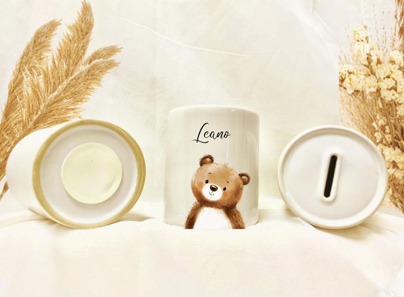 May include: White ceramic piggy bank with a cute brown bear illustration and the name "Leano" in script. The bank is cylindrical with a slot for coins on top and a removable base. Decorative dried plants are in the background.
