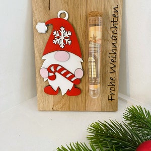 May include: Wooden Christmas decoration featuring a gnome with a red hat and candy cane. A clear tube with a cork stopper contains rolled-up paper money. The words "Frohe Weihnachten" are etched on the wood.
