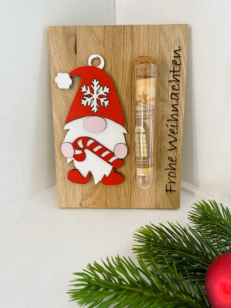 May include: Wooden Christmas decoration featuring a gnome with a red hat and candy cane. A clear tube with a cork stopper contains rolled-up paper money. The words "Frohe Weihnachten" are etched on the wood.