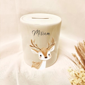May include: A cream-colored ceramic bank with a slot on top. The bank features a cartoon deer illustration in brown and white, with the name "Miriam" in black script. The bank is cylindrical and sits on a white surface.
